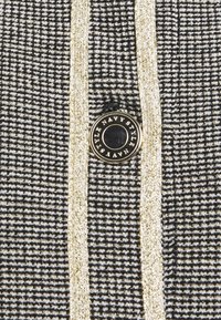 Tela de pata de gallo en blanco y negro con detalles en dorado. Presenta un botón negro prominente con una inscripción dorada. Superficie texturizada y diseño estructurado.