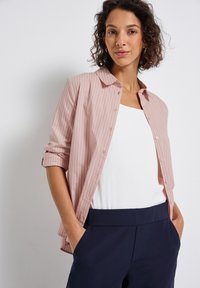 Donna con capelli ricci che indossa una camicia a righe verticali rosa chiaro sopra una maglietta bianca e pantaloni blu scuro, con le mani nelle tasche.