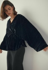 Black velvet wrap top with long, wide sleeves and a knotted detail at the waist, paired with a form-fitting black skirt. Smooth texture.