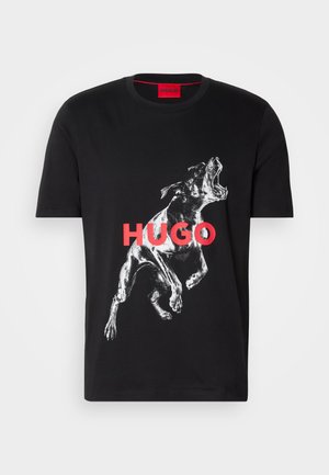 Melns kokvilnas T-krekls ar baltu grafiku, kurā ir suns vidējā zīlē, un vārds "HUGO" treknām sarkanām burtiem krūšu rajonā.