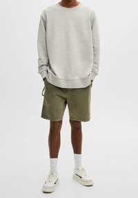 Felpa grigia con scollatura a girocollo, maniche lunghe e polsini a coste, abbinata a pantaloni corti verde oliva. Sneakers con base bianca e dettagli neri.