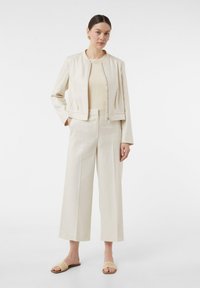 Beige cropped jack met een ronde hals, gecombineerd met een bijpassend beige top en wijde broek. De sandalen hebben een gevlochten detail.
