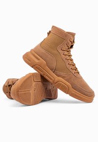 Brązowe wysokie buty zamszowe z tkaniny, wyposażone w gumowe podeszwy z teksturowanym wzorem antypoślizgowym i sznurowanym designem.
