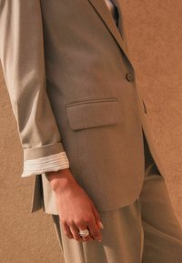 Light gray tailored suit with vertical stripes; features a single button, flap pocket, and contrasting white striped cuffs. Jewelry includes layered rings.