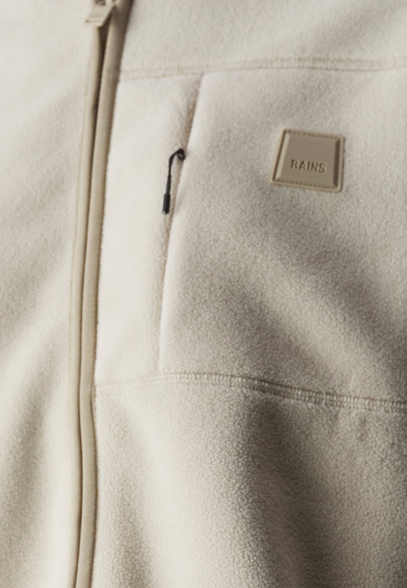 Beige fleecejack met een ritssluiting, voorzien van een borstzak en een klein vierkant logo-embleem. Zachte textuur en contrasterende bruine accenten.