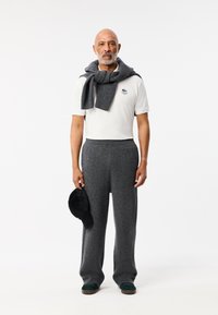 Šedé pletené kalhoty, bílé polo tričko s logem, šedý šátek přes ramena, černá kšiltovka v ruce, tmavě zelené boty. Jednoduchý, uvolněný outfit bez vzorů.