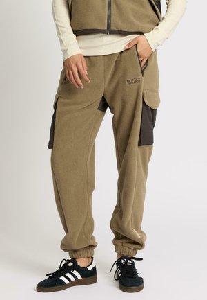 Personne portant un pantalon cargo en polaire beige avec des poches latérales, des baskets noires et blanches, et un gilet en polaire assorti, se tenant avec une main dans la poche.
