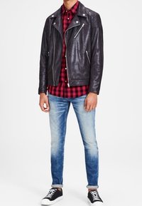 Chaqueta de biker de cuero negro con cremalleras plateadas, llevada sobre una camisa a cuadros rojos y negros. Unos vaqueros azules y unas zapatillas negras completan el conjunto.