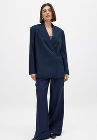 Femme aux cheveux bruns courts portant un blazer oversize bleu foncé et un pantalon large assorti, se tenant contre un fond clair uni.