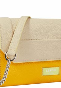 Sac à main en cuir beige et jaune avec une finition texturée, une chaîne en bandoulière et une petite étiquette de marque. Dispose d'une fermeture à rabat et de couleurs contrastantes.