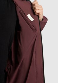 Burgundy jacket with a smooth texture, featuring a zip closure and an inside pocket with a label. Partially open, revealing interior details.