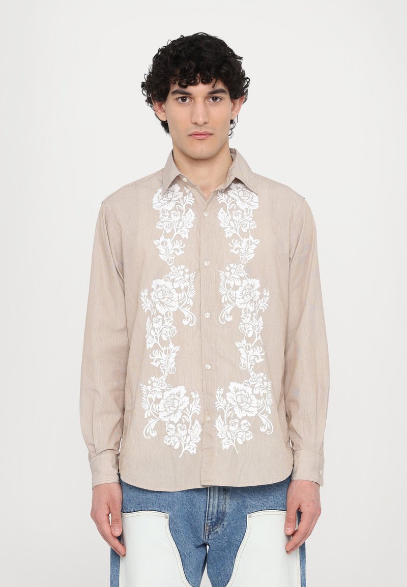 Camicia a maniche lunghe marrone chiaro con ricami floreali bianchi sulla parte anteriore, dotata di colletto a punta e maniche con polsini abbottonati.