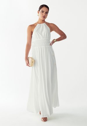 Žena má na sebe biele dlhé šaty bez rukávov s halter výstrihom, v jednej ruke drží béžovú kabelku, zlaté kruhové náušnice, náramok a sandále.