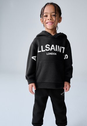 Hymyilevä lapsi, jolla on punotut hiukset, pukeutuneena mustaan AllSaints London -huppariin ja mustiin housuihin, seisoo yksivärisen vaalean taustan edessä.