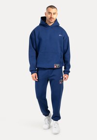 Felpa blu navy con tasca frontale e logo, abbinata a pantaloni della tuta coordinati con etichette in tessuto e sneakers bianche.