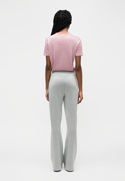 Personne aux cheveux longs tressés portant un haut rose à manches courtes et un pantalon large taille haute argenté, debout de dos sur un fond uni.