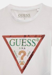 Valkoinen orgaanisesta puuvillasta valmistettu t-paita, jossa on suuri, vihreästä, punaisesta ja kullasta koostuva paljettitrikooli, jonka tekstit ovat "GUESS U.S.A." ja "WASHED JEANS."