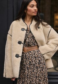 Beige shearlingjacka med svarta detaljer, med knäppningar i togglestil. Lagd över en blommig klänning i bruna nyanser, som accentueras av ett flätat brunt bälte.
