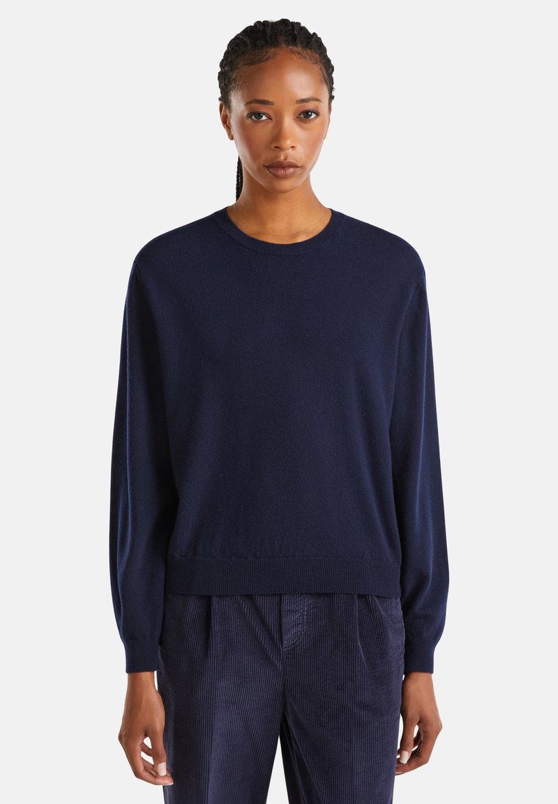 Femme portant un pull à manches longues bleu marine et un pantalon en velours côtelé bleu foncé, se tenant devant un fond blanc uni.