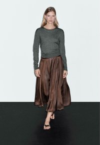 Vrouw die op een zwarte vloer loopt, gekleed in een donkergrijze langemouwen top, een bruin satijnen geplooide rok, zwarte sandalen en zilveren oorbellen tegen een witte muur.