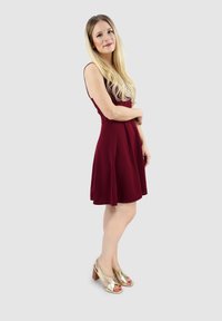 Bordeaux mouwloze jurk met een uitlopende rok; model draagt gouden hoge hakken. Glad textiel, knie-lange ontwerp. Neutrale achtergrond.