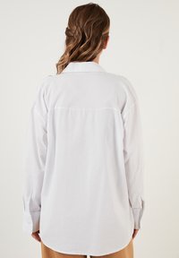 White long-sleeve shirt with a classic collar, featuring a loose fit, side slits, and a curved hem. The material appears smooth and lightweight.