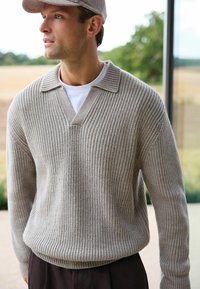 Lys grå, ribbet striktrøje med V-hals og kort, struktureret krave. Bæres over en hvid T-shirt og parret med mørke bukser.
