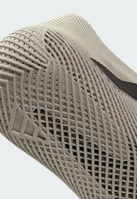 Skoens overdel i lyst gråt materiale har en tekstureret overflade med geometriske mønstre og ventilationshuller; sort brandingdetalje er synlig.