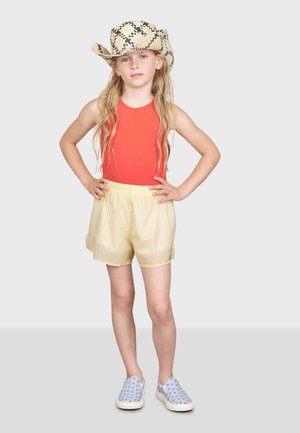 Röd ärmlös bodysuit, gula shorts och blåa slip-on skor. Bär en halm cowboyhatt med svarta mönster. Långt vågigt hår.