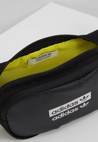 Riñonera negra de Adidas con logo blanco, sin cerrar para mostrar el brillante forro interior amarillo y la etiqueta de la marca.