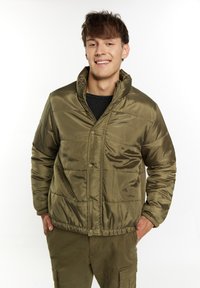 Giacca puffer verde oliva con colletto alto, bottoni a pressione e texture trapuntata, abbinata a un top in rete nera e pantaloni coordinati.