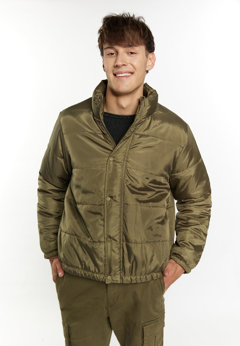 Giacca puffer verde oliva con colletto alto, bottoni a pressione e texture trapuntata, abbinata a un top in rete nera e pantaloni coordinati.