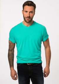 T-shirt turchese a scollo a V con maniche arrotolate, realizzato in tessuto morbido. Il modello indossa jeans scuri, mostrando un tatuaggio sul braccio sinistro.