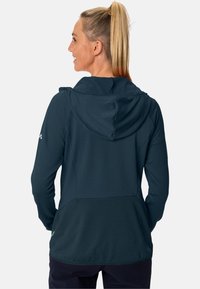 Giacca cappuccio blu scuro in tessuto testurizzato, dal design aderente, con tasche posteriori. Presenta un cappuccio regolabile con cordino e un dettaglio con logo sulla manica.