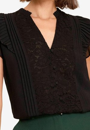 Close-up van een vrouw die een zwart overhemd draagt met een kanten voorkant, knopen bedekt met stof, en geplooide korte mouwen, ingestopt in een groene broek.