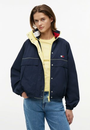 Młoda kobieta ubrana w granatową bluzę na zamek z żółtą koszulką i jasnoniebieskimi dżinsami, stojąca z jedną ręką w kieszeni.