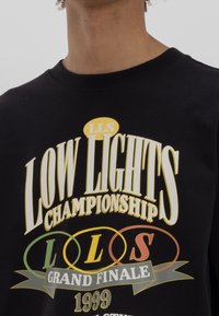 Černé bavlněné tričko s výrazným bílým a žlutým textem. Grafika zahrnuje "LOW LIGHTS CHAMPIONSHIP" a "GRAND FINALE 1999" s barevnými kruhy.