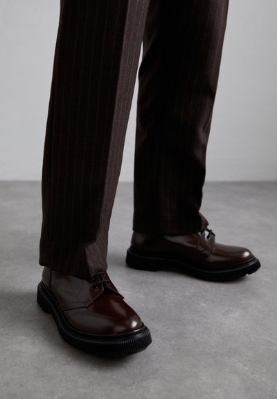 Braune Leder Schnürschuhe mit glänzender Oberfläche, schwarzer Gummisohle und dezenten Nähten. Getragen mit braunen Angabenhosen mit Nadelstreifen.