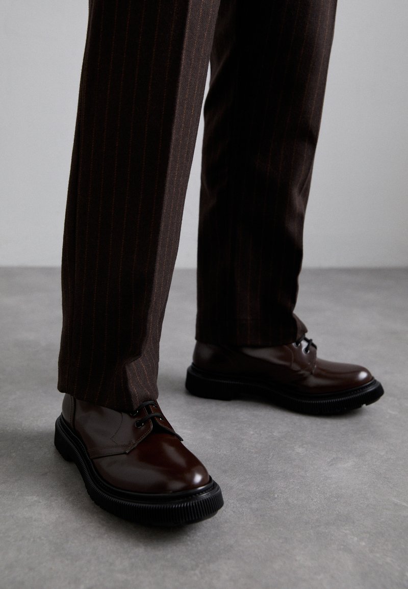 Chaussures en cuir marron à lacets avec une finition brillante, semelle en caoutchouc noire et coutures discrètes. Portées avec un pantalon marron à rayures.