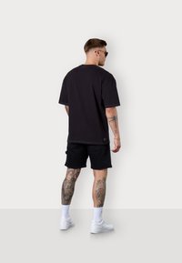 Black short-sleeve t-shirt features a relaxed fit, white contrast stitching, paired with black shorts and white sneakers. Visible tattoos on legs.