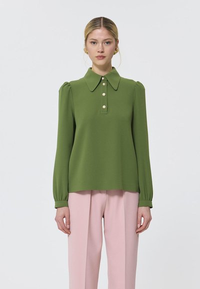 Blouse verte avec un col et une patte de boutonnage à quatre boutons, avec des manches bouffantes et une texture lisse, associée à un pantalon rose.