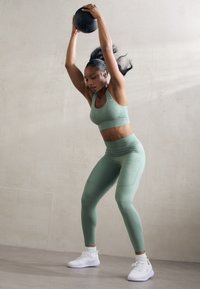 Conjunto deportivo de tela verde menta, sujetador deportivo ajustado y mallas, sosteniendo una pelota medicinal negra, con zapatillas deportivas blancas, realizando un ejercicio.