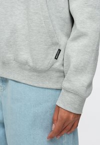 Grå genser med teksturert stoff, side lomme og en svart etikett. Mansjetten er tettsittende, og den er kombinert med lyseblå jeans.