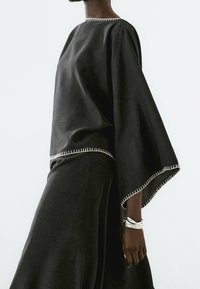 Haut noir ample avec larges manches, présentant des coutures blanches le long de l'encolure et de l'ourlet. Texture lisse, design minimaliste.