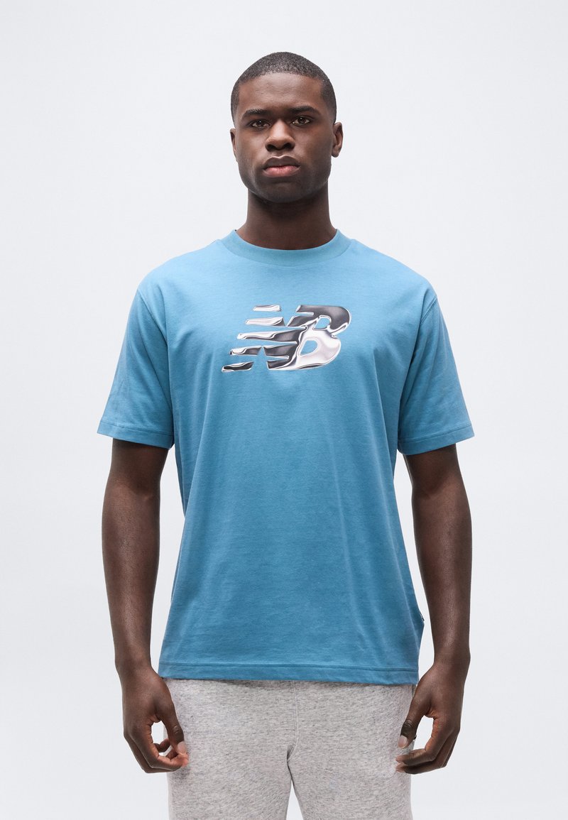 Νεαρός άνδρας που φοράει μπλε μπλούζα με λαμπερό ασημένιο λογότυπο "NB" και ανοιχτό γκρι παντελόνι, στέκεται μπροστά σε ένα απλό λευκό φόντο.