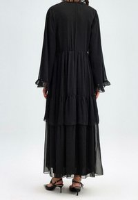 Robe maxi noire à volants avec des manches longues et des ourlets ruffled. Confectionnée dans un tissu léger, elle présente un col haut et des couches transparentes.