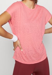 Różowa koszulka z krótkim rękawem z delikatnym wzorem zebry, wykonana z lekkiego materiału o luźnym kroju. Noszona z bordowymi legginsami.