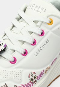 Λευκό sneaker Skechers με ροζ και πορτοκαλί τρυπούλες, λευκές κορδόνες, σχεδιασμούς με λουλούδια και κρανία στη σόλα, και σήμα "SKECHERS".