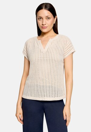 Kvinde iført beige kortærmet striktop med udskæring i halsudskæringen og navy bukser, stående mod en ensfarvet lys baggrund.