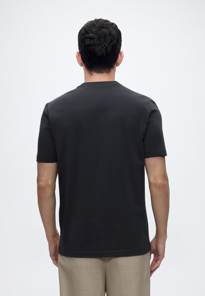 Homme aux cheveux courts et foncés portant un t-shirt noir uni à manches courtes et un pantalon beige, vu de dos sur fond blanc.